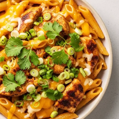 A close-up of a bowl of creamy BBQ chicken pasta with penne, topped with blackened chicken pieces, melted cheddar cheese, fresh cilantro, and sliced green onions.
