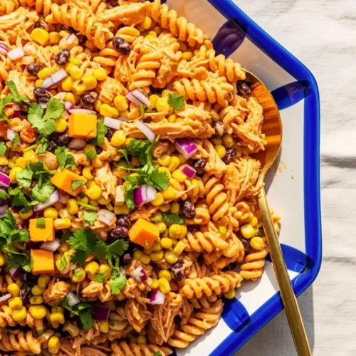 An overhead shot of a Southwestern BBQ chicken pasta salad in a decorative serving dish, featuring rotini pasta, shredded chicken, black beans, corn, diced cheddar cheese, and red onion, garnished with fresh cilantro.
