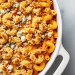 A close-up of a baked buffalo chicken mac and cheese casserole, featuring elbow macaroni and shredded chicken in a creamy cheese sauce, finished with a golden-brown breadcrumb topping and blue cheese crumbles.
