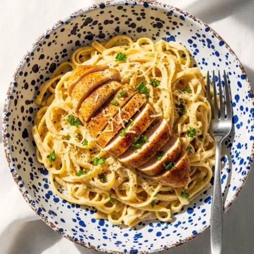 An overhead view of a bowl of creamy chicken Fettuccine Alfredo, featuring long pasta noodles tossed in a rich Parmesan cream sauce, topped with sliced seasoned chicken and fresh parsley.