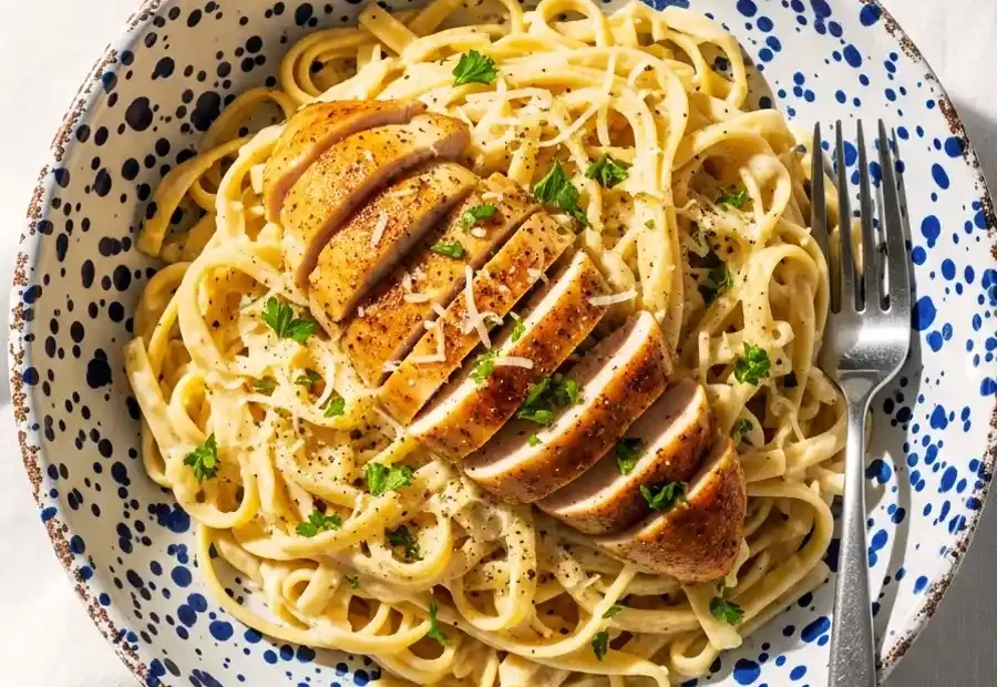 An overhead view of a bowl of creamy chicken Fettuccine Alfredo, featuring long pasta noodles tossed in a rich Parmesan cream sauce, topped with sliced seasoned chicken and fresh parsley.