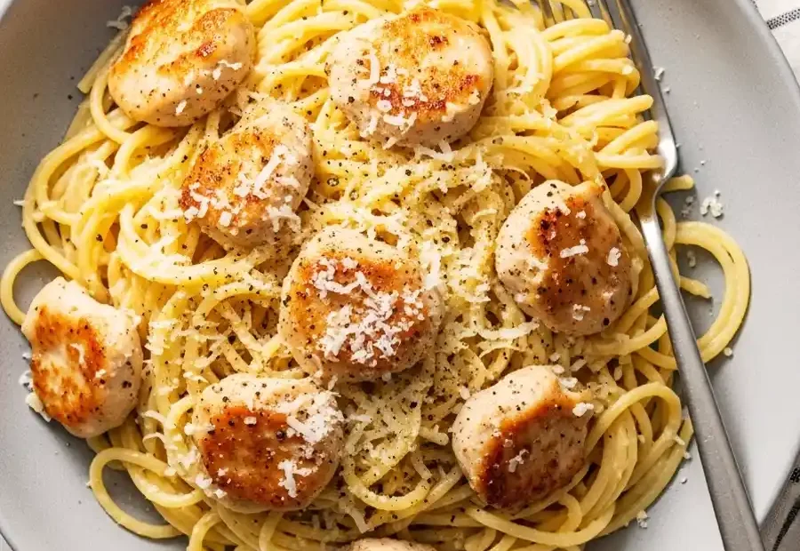 An overhead shot of a bowl of chicken carbonara, featuring spaghetti tossed in a creamy egg and Parmesan sauce, topped with pan-seared chicken medallions and black pepper.