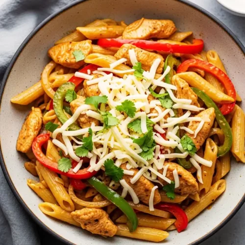 A bowl of creamy chicken fajita pasta, featuring penne tossed with seasoned chicken strips and sautéed red and green bell peppers in a zesty chili-lime sauce, topped with shredded Monterey Jack cheese and fresh cilantro.
