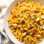 An overhead view of a large white bowl filled with creamy homemade chicken mac and cheese, featuring elbow macaroni in a rich cheddar sauce, topped with diced seared chicken, and garnished with black pepper and parsley.