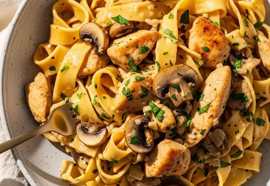 A white bowl of creamy Chicken Marsala pasta, featuring fettuccine noodles tossed with sautéed chicken and cremini mushrooms in a rich and savory Marsala sauce.