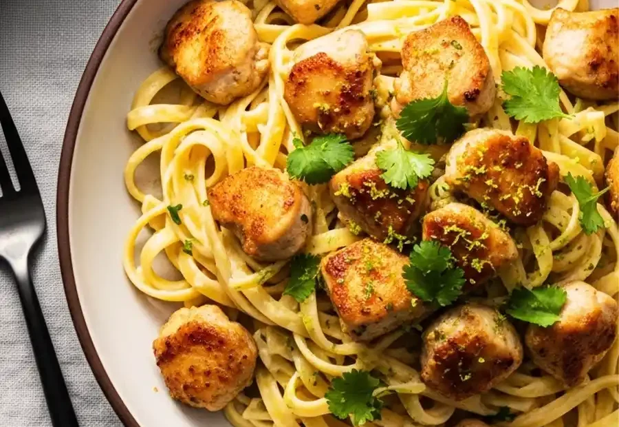 A close-up of a bowl of cilantro lime chicken pasta, featuring linguine noodles tossed in a zesty cream sauce, and topped with pan-seared chicken bites, fresh cilantro, and lime zest.