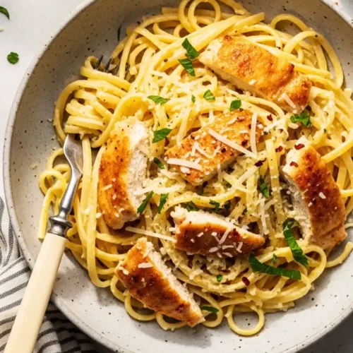 A bowl of creamy lemon chicken pasta, featuring linguine in a light lemon-parmesan cream sauce, topped with sliced golden-brown chicken, fresh parsley, and red pepper flakes.