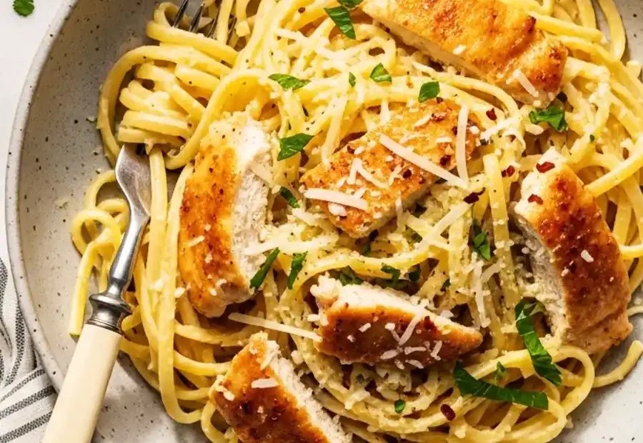 A bowl of creamy lemon chicken pasta, featuring linguine in a light lemon-parmesan cream sauce, topped with sliced golden-brown chicken, fresh parsley, and red pepper flakes.