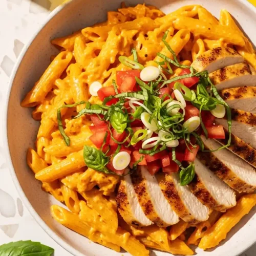 A bowl of copycat Firebirds Chicken Pasta, with penne in a spicy Asiago cream sauce, topped with sliced grilled chicken, and a fresh garnish of diced tomatoes, basil, and green onions.