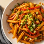 A white bowl filled with spicy Rasta Pasta, featuring penne tossed with jerk chicken, sautéed red and green bell peppers, and a creamy Caribbean-style sauce, garnished with fresh green onions.