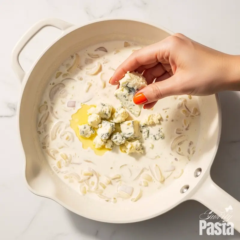 Overhead view of crumbled gorgonzola cheese melting into a simmering cream sauce in a white skillet.