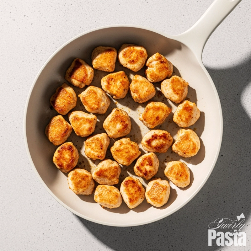 Overhead view of bite-sized chicken pieces being seared to a golden brown in a matte white ceramic skillet.