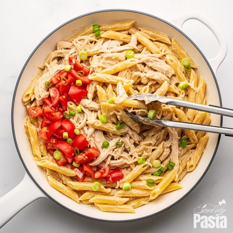 Overhead view of cooked penne pasta, chicken, and tomatoes being tossed with Asiago cream sauce in a large white skillet.
